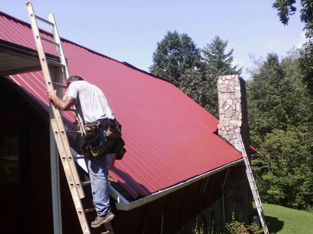 Roofer on a ladder beside a red metal roof and stone chimney — Soffit & Fascia Repair by Roofing Brothers in Jamestown